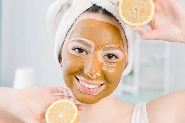 Beautiful young woman with facial mud mask and halfs of lemon on light background in spa center — Stock Photo, Image