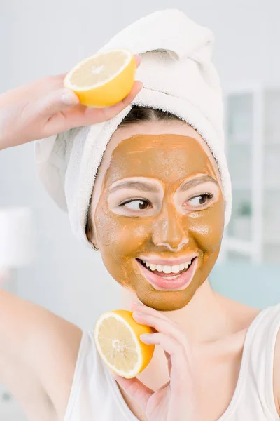 Beauty Skin Care Concept. Bella donna caucasica in asciugamano bianco con maschera facciale marrone fango sul viso tiene agrumi sulla mano su sfondo chiaro. Procedure di Stazione termale e maschera su pelle — Foto Stock