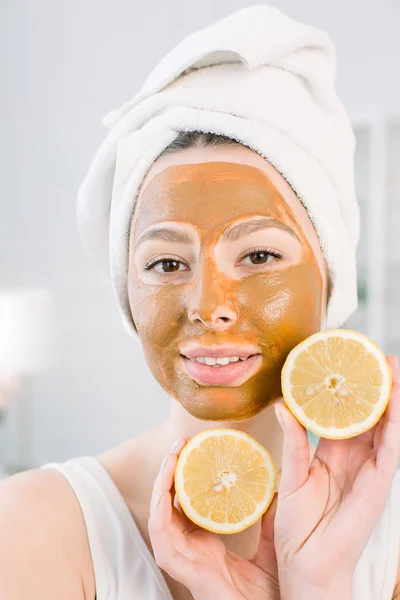 Beauty Skin Pielęgnacja koncepcja. Ładna kaukaski kobieta w białym ręczniku z brązowym błocie twarzy maska na twarzy trzyma owoców cytrusowych na rękę na jasnym tle. Zabiegi spa i maska na skórze — Zdjęcie stockowe