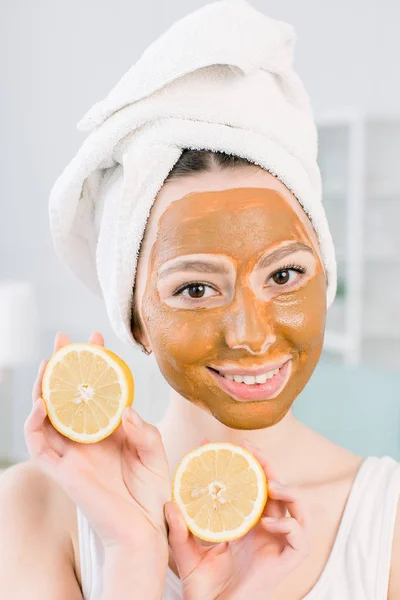 Indah, tersenyum gadis di handuk putih dan lumpur cokelat masker wajah bersenang-senang dengan dua bagian dari lemon, tembakan dalam ruangan di latar belakang putih — Stok Foto