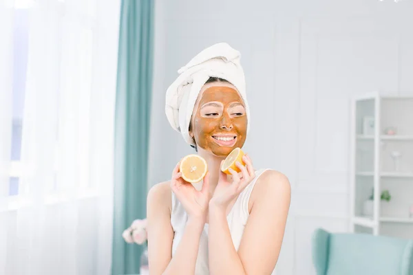 Belleza procedimientos cuidado de la piel concepto. Mujer joven con mascarilla de barro facial marrón en su cara en el baño, con toalla blanca en la cabeza, sosteniendo mitades de limón —  Fotos de Stock