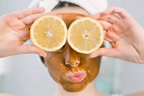 Menina bonita nova com máscara de lama marrom no rosto segurando metades de frutas de limão, cobrindo os olhos. Menina adolescente cuidando de sua pele. Tratamento da beleza . — Fotografia de Stock