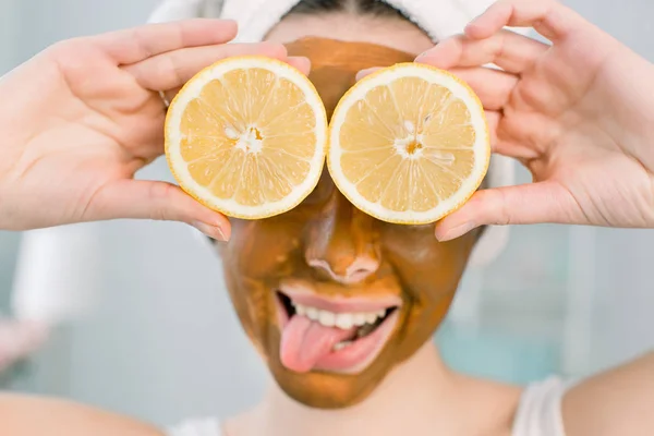 Menina bonita nova com máscara de lama marrom no rosto segurando metades de frutas de limão, cobrindo os olhos. Menina adolescente cuidando de sua pele. Tratamento da beleza . — Fotografia de Stock