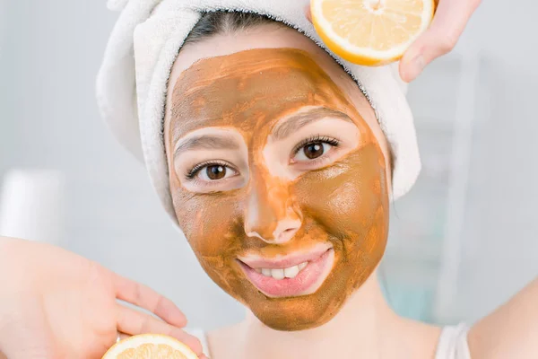 Indah, tersenyum gadis di handuk putih dan lumpur cokelat masker wajah bersenang-senang dengan dua bagian dari lemon, tembakan dalam ruangan di latar belakang putih — Stok Foto