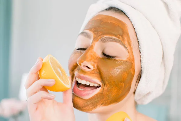 Wanita muda yang menarik dengan handuk putih di kepala dan dengan masker coklat wajah lumpur menggigit lemon dan tersenyum. Kecantikan, spa, perawatan kulit dan tubuh . — Stok Foto