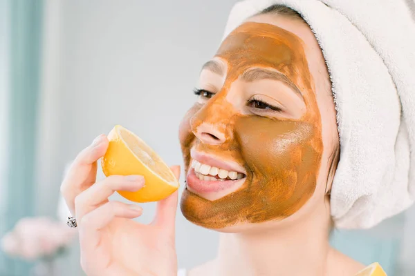 Wanita muda yang menarik dengan handuk putih di kepala dan dengan masker coklat wajah lumpur menggigit lemon dan tersenyum. Kecantikan, spa, perawatan kulit dan tubuh . — Stok Foto