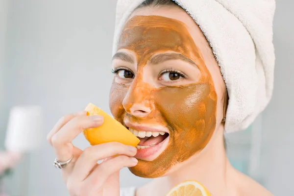 Jovem atraente com toalha branca na cabeça e com máscara marrom facial de lama morde um limão e sorri. Beleza, spa, cuidados com a pele e o corpo . — Fotografia de Stock