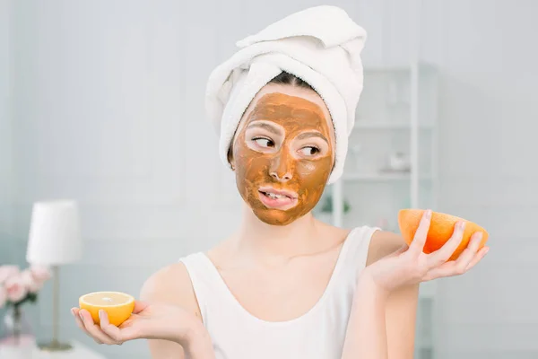 Mujer hermosa divertida sosteniendo rodajas de limón y pomelo. Foto de niña en toalla blanca con máscara facial de barro marrón, que tiene procedimientos de spa . —  Fotos de Stock