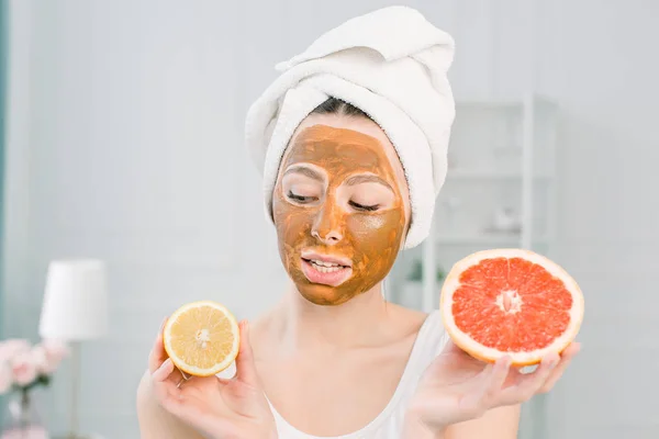 Funny beautiful woman holding lemon and grapefruit slices. Photo of girl in white towel with brown mud facial mask, having spa procedures.