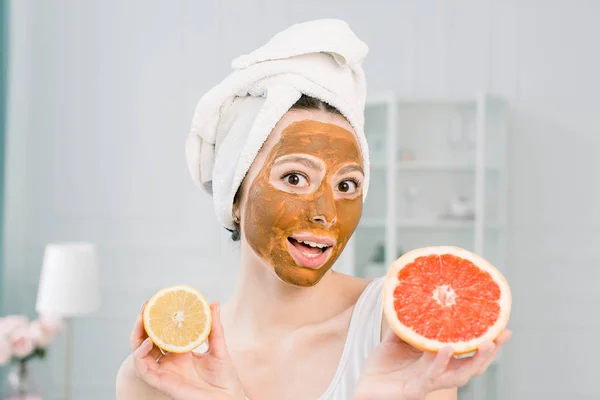 Mujer hermosa divertida sosteniendo rodajas de limón y pomelo. Foto de niña en toalla blanca con máscara facial de barro marrón, que tiene procedimientos de spa . —  Fotos de Stock