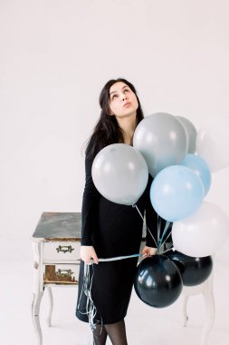 Beyaz bir arka plan üzerinde balonlar ile güzel genç kadın, vintage beyaz masa nın yanında duran ve yukarı bakıyor. Doğum günü ve parti. Festivalde siyah elbiseli kız