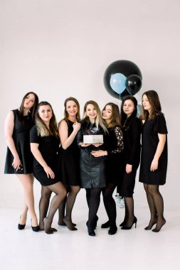 Portrait of beautiful bridesmaids in black dresses with attractive bride-to-be in the middle holding delicious cake. Background is decorated with air balloons. clipart
