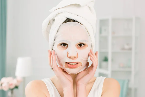 Gadis muda dengan masker wajah melihat kamera di atas latar belakang cahaya. Prosedur Kosmetik Kecantikan spa dan kosmetologi . — Stok Foto