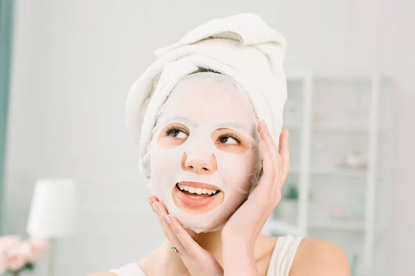 Mulher bonita nova com toalha branca na cabeça, aplicando em uma máscara facial, conceito de cuidados com a pele. Spa, saúde. Mulher com máscara purificadora em seu rosto — Fotografia de Stock