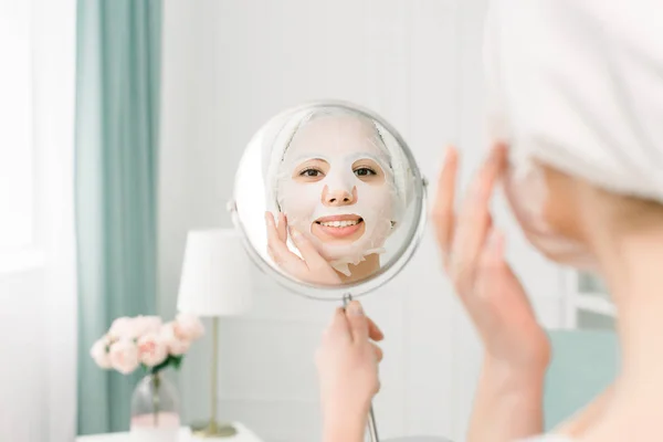 Perawatan wajah dan perawatan kecantikan. Wanita dengan lembaran masker pelembab di wajahnya dan handuk putih di kepala, melihat cermin . — Stok Foto