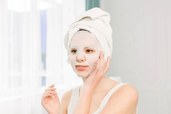 Szoros portré fiatal csinos nőről, aki papírlepedő maszkot visel az arcára. Kozmetikai eljárás. Szépségfürdő és kozmetika. — Stock Fotó