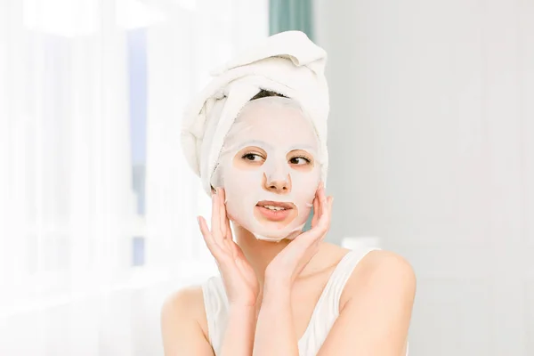 Красотка. Крупный план улыбающейся молодой женщины со свежим натуральным макияжем, наносящим маску из текстильного листа. Портрет привлекательной счастливой девушки в белой косметической маске . — стоковое фото