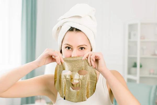 Mladá hezká žena v bílém ručníku po koupeli se zlatou papírovou maskou. Žena dostává zlatou obličejovou masku. — Stock fotografie