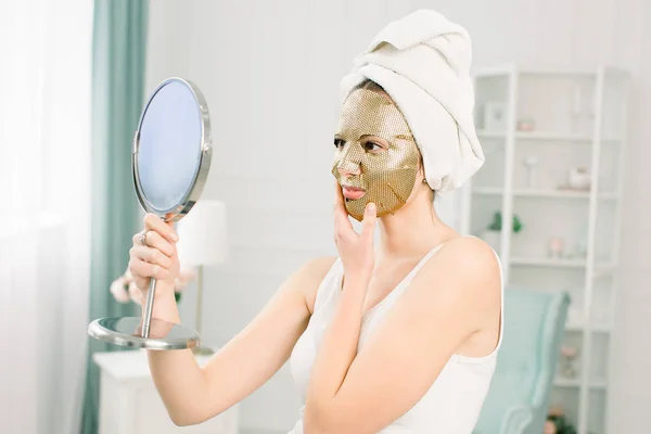 Perawatan wajah dan perawatan kecantikan. Wanita dengan lembaran pelembab masker emas di wajahnya dan handuk putih di kepala, melihat cermin . — Stok Foto