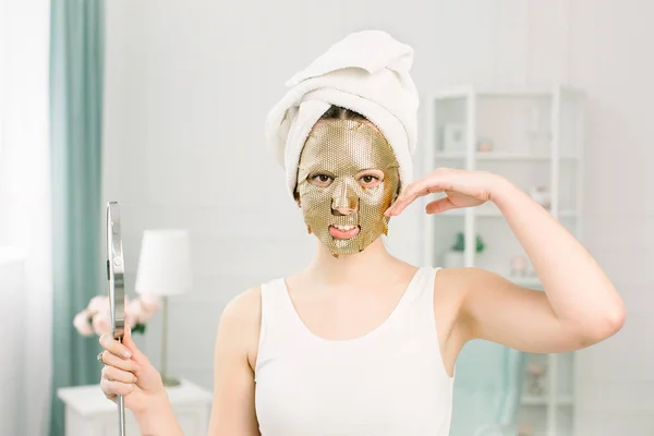 Gouden masker cosmetische procedure in de schoonheidssalon. Aantrekkelijk sexy meisje met witte handdoek aanraken gezicht en gouden masker op gezicht, het houden van een spiegel. — Stockfoto