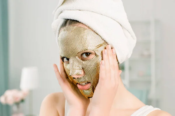 Złota Maska Kobiet. Piękna młoda kobieta w białym ręczniku na głowie ze Złotą Skórą Kosmetykiem, patrząca w kamerę. Pielęgnacja i pielęgnacja skóry piękna — Zdjęcie stockowe
