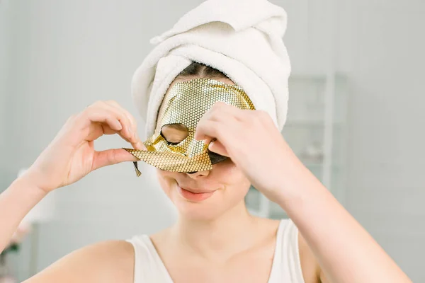 Maska ženské tváře. Detailní záběr krásná usmívající se dívka v bílém ručníku sundání kosmetické prostěradlo zlatá maska ze zdravé kůže. — Stock fotografie