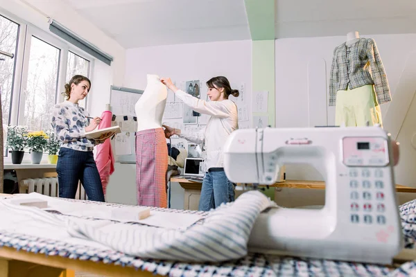 Две портнихи или швея, выполняющие измерения манекена с помощью измерительной ленты в студии дизайна одежды, женщина-дизайнер, работающая с эскизами в мастерской, концепция одежды и шитья — стоковое фото