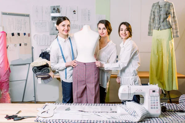 Três estilistas felizes trabalhando com tecido, esboços de roupas, materiais de medição em manequim no estúdio cheio de ferramentas e equipamentos de alfaiataria — Fotografia de Stock