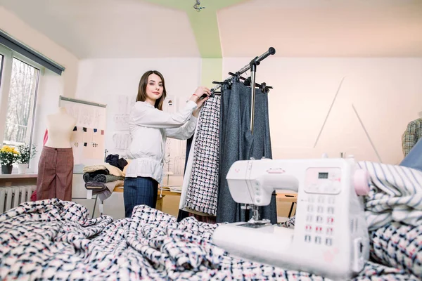 Female clothes designer working with new woman pants collection in cozy workshop studio. Fashion designer, dressmaker standing near clothes rack, touching, holding in hands fashionable handmade pants