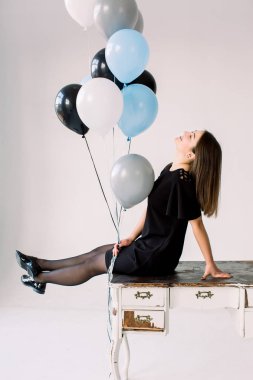 Siyah, beyaz, mavi ve gri balonlar ile poz siyah elbise zarif ince doğum günü kız. Beyaz arka plan üzerinde masada oturan sevimli gülen doğum günü kız tam uzunlukta portre