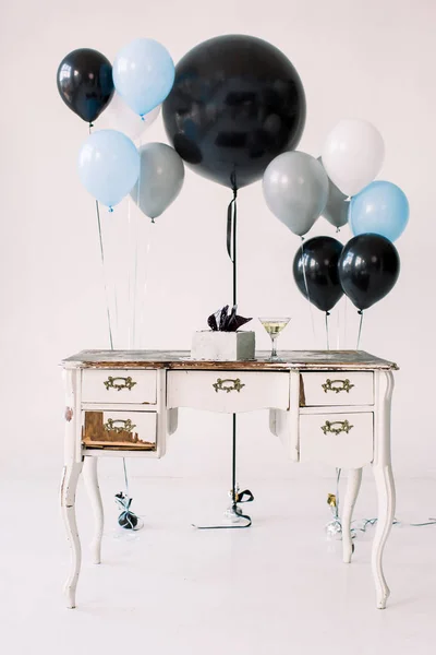 Vintage white wooden table with closets, birthday cake, martini glass and black, blue and gray air balloons, isolated on white background. Holiday, birthday party concept