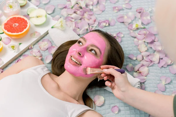 Cosmetologist smears cosmetic facial mask on the face of the pretty young woman in the spa salon. Face mask, spa beauty treatment, skincare.