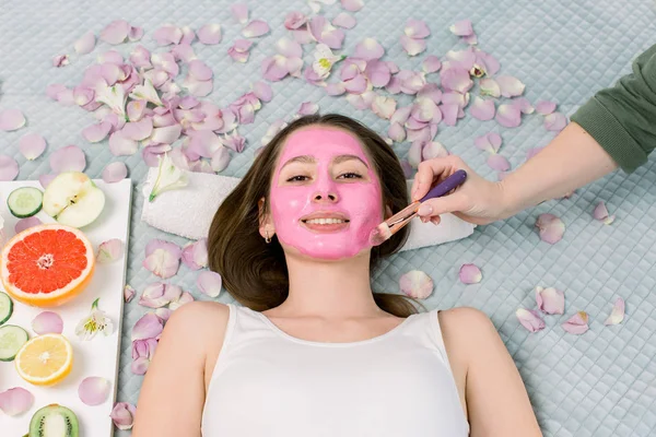 Maschera viso, cura della pelle, procedure termali. Bella ragazza al centro benessere, sdraiata sul letto con petali di fiori e frutta fresca. Cosmetico donna applicando maschera facciale rosa — Foto Stock