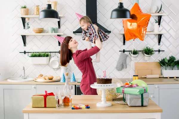 Boldog anya tartja a lányát a kezét a háttérben a világos konyha, születésnapi torta, jelenlegi dobozok, léggömbök. Szülinapos bébi — Stock Fotó