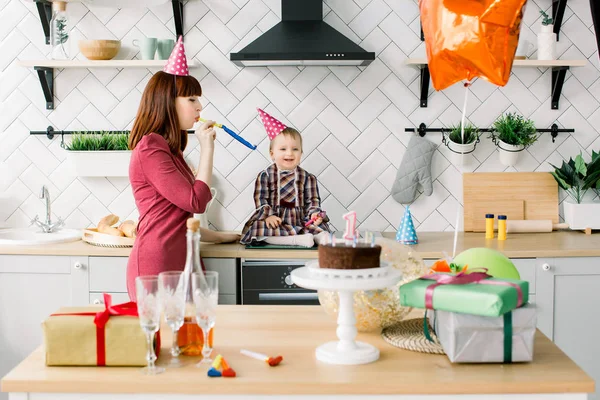 Mother celebrating first birthday of her baby girl at home kitchen. Baby girl smiling and having fun while her pretty mother blowing into party horn