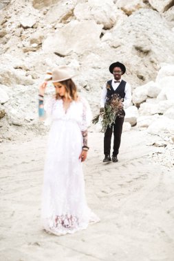 Açık kahverengi şapka lı ve boho elbiseli genç bir kadın gelin çöl kanyonunun arka planında duruyor. Arka planda takım elbiseli Genç Afrikalı adam düğün buketi tutar, adam odaklanmak