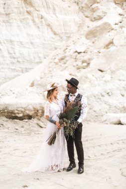 Çöl Kanyonu'nda gelin ve damadın ilk buluşması. Rustik boho düğün töreni açık havada. Beyaz elbiseli güzel Beyaz gelin ve siyah takım elbise ve şapkalı Afrikalı damat