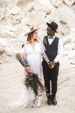 Beautiful boho-style multi racial couple newlyweds walking in nature desert canyon, summer day. African man and Caucasian woman looking each other and holding hands clipart