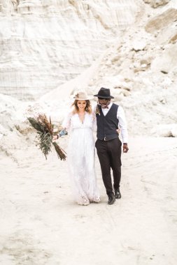 Çöl kanyonunda birlikte yürüyen ve birbirlerine şefkat ve sevgiyle bakan yeni evliler kucaklaşıyor ve el ele tutuşuyorlar. Kanyon arka planda Boho ve rustik şık düğün