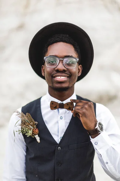 Close-up portret van knappe glimlachende Afro Amerikaanse bruidegom in zwarte hoed, wit overhemd en zwart zwart vest poseren in Canyon tegen prachtige woestijnlandschap bij zonsondergang. — Stockfoto