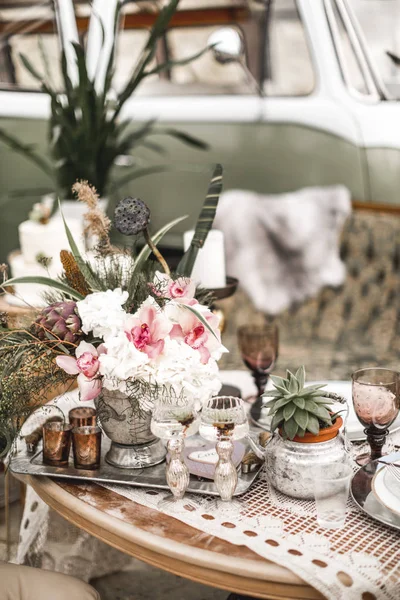 Beautiful glasses, candles, flowers stand on a fruitful decorated table in rustic boho style. A large green sofa and hippy bus on the background. Summer desert atmosphere. Wedding party — Stock Photo, Image