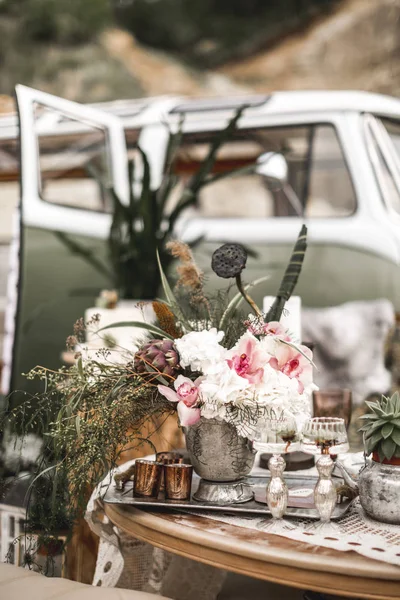 Schöne Gläser, Kerzen, Blumen stehen auf einem fruchtbar dekorierten Tisch im rustikalen Boho-Stil. Im Hintergrund ein großes grünes Sofa und ein Hippiebus. sommerliche Wüstenatmosphäre. Hochzeitsgesellschaft — Stockfoto