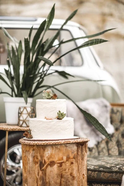 Elegante weiße zweistöckige Hochzeitstorte mit Blumen und Sukkulenten auf dem Holzbalken auf dem Hintergrund von Dekorationen im rustikalen Stil — Stockfoto