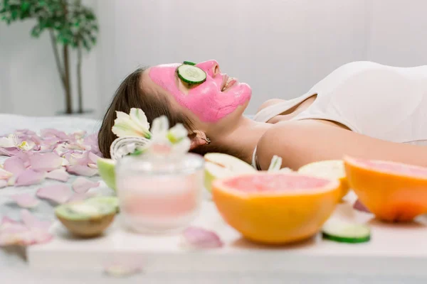 Hermosa joven mujer está recibiendo máscara de arcilla facial en el spa, acostado con pepinos en los ojos. Vista lateral. Cuidado de la piel, concepto de cosmética natural —  Fotos de Stock