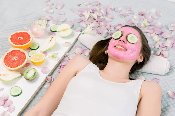 Schöne junge Frau mit rosa alginatfarbener Gesichtsmaske auf dem Gesicht und Scheiben frischer Gurken auf dem Bett liegend im Wellnessbereich. Hautpflege und -behandlung, Wellness, natürliche Schönheit und kosmetologisches Konzept — Stockfoto