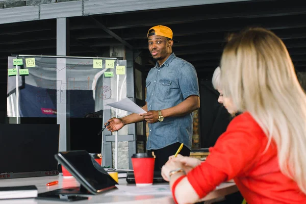 Les jeunes employés de bureau assis à la table tandis que le collègue africain masculin montrant des idées sur le mur de verre lors de la réunion, groupe d'employés prospères appréciant le processus de travail, et le remue-méninges — Photo