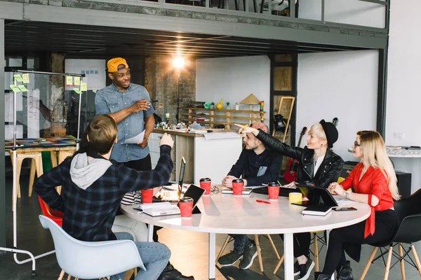 Nonchalant klädd multirasistiska manliga och kvinnliga studenter grupp ha kul samtidigt lära sig tillsammans i Coworking rymden, hipster afrikansk kille som använder glas kontors kort för att göra tal medan brainstorming — Stockfoto