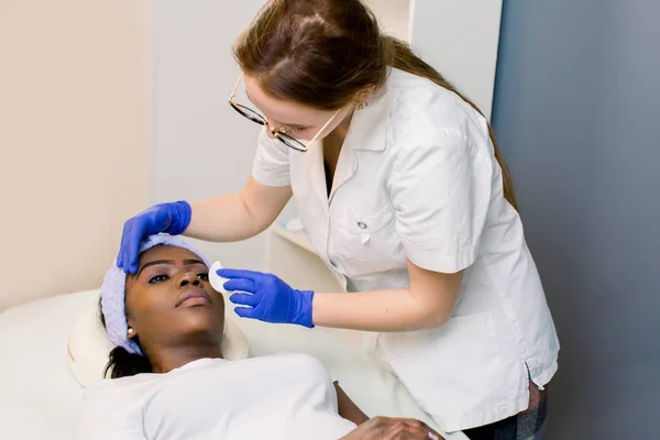 Cosmetologa donna al lavoro. Estetista pulizia della pelle sulle palpebre attenti. Giovane donna africana sdraiata sul divano in camera. Cosmetologia e cura della pelle — Foto Stock