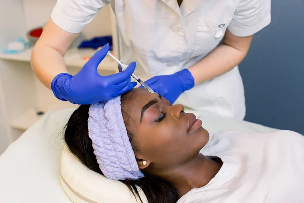 Nahaufnahme der Hände einer Kosmetikerin, die einem jungen afrikanischen Mädchen Botox in die Stirn spritzt. — Stockfoto