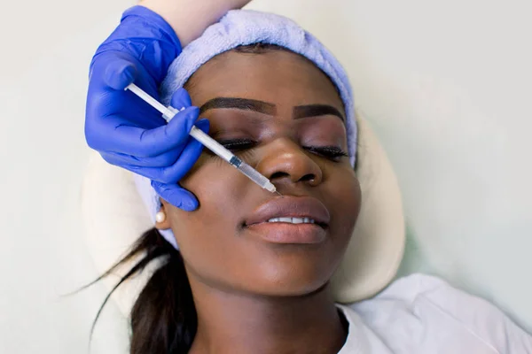 Beautiful young African woman gets cosmetic injection on her lips, isolated over light background. Doctor is making an injection in lips. Beauty Treatment.